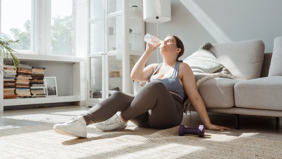 Woman sitting on living room floor, drinking water after workout, enjoying relaxation.